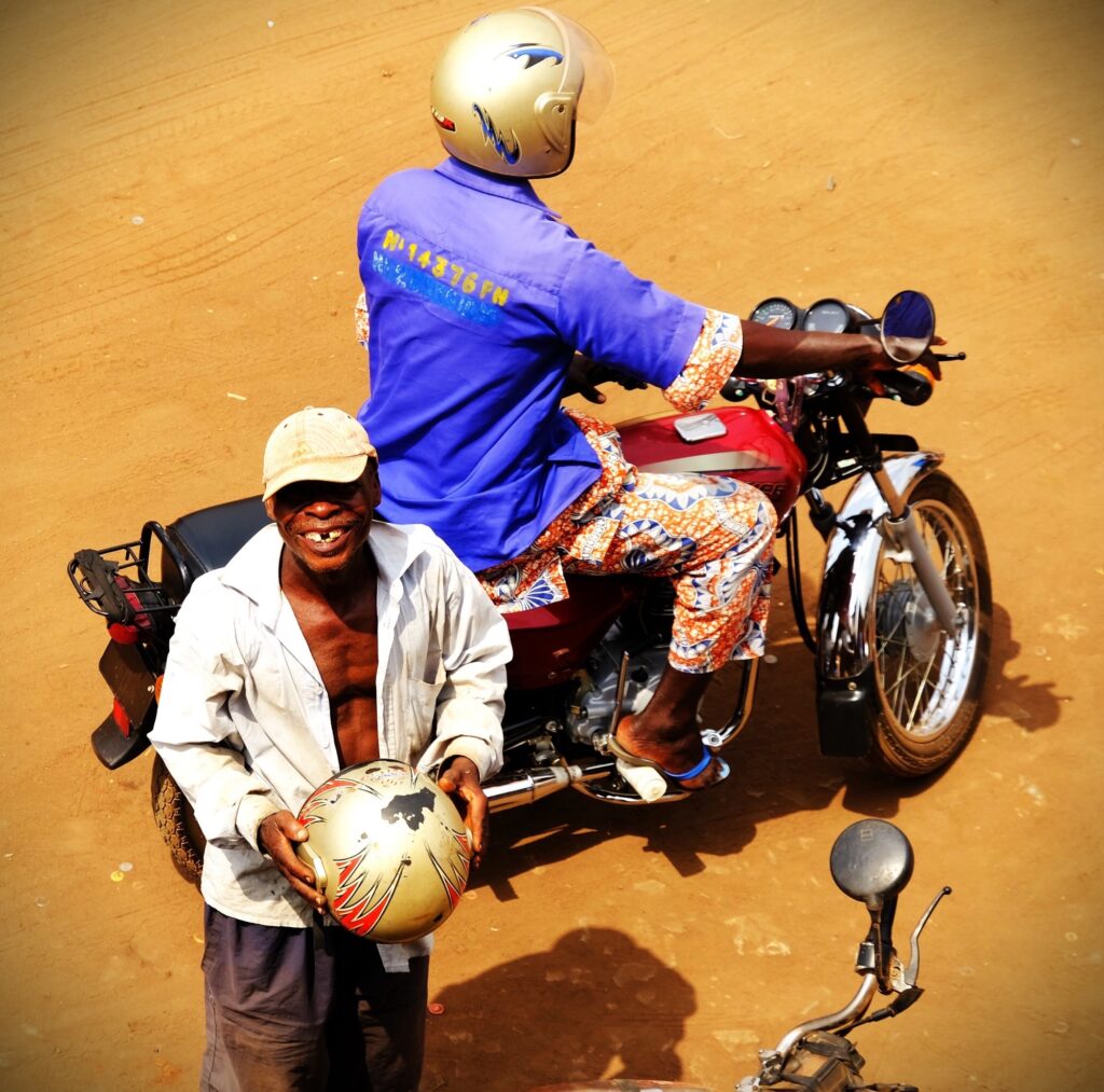 un homme souriant avec les dents du bonheur, derrière lui un moto taxi (zem ou kekeno) sur sa moto en claquette